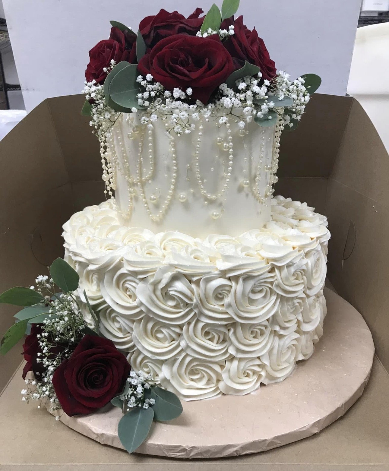Two-tier white buttercream wedding cake with piped rosettes, strands of pearls and fresh red roses with baby's breath