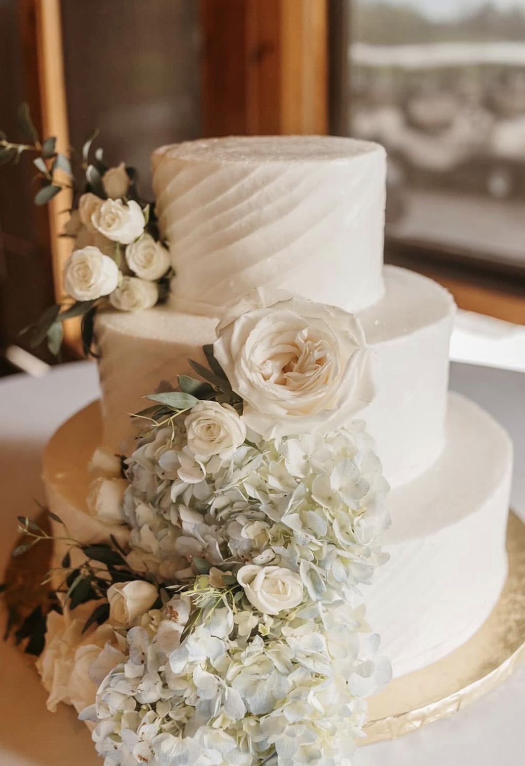 Three-tier white buttercream wedding cake with textured icing and a cascade of white roses, hydrangeas and eucalyptus